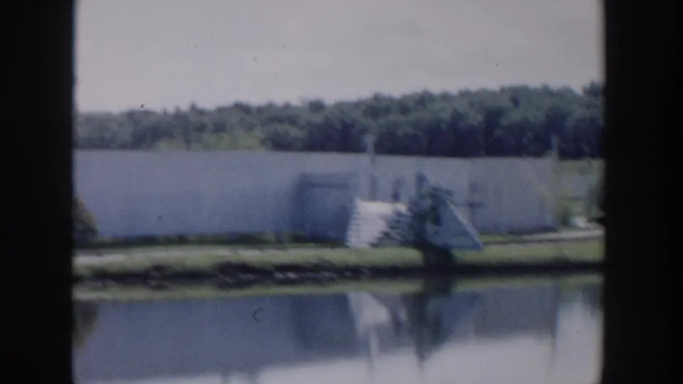 1954 Wisconsin Dells frame showing a pale lakeside wall, steps or dock area, people, trees, and calm reflective water.