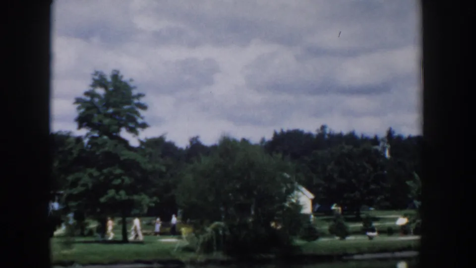 1954 Wisconsin Dells 8mm frame showing a green park lawn, trees, distant visitors, and a cloudy sky from a moving viewpoint.