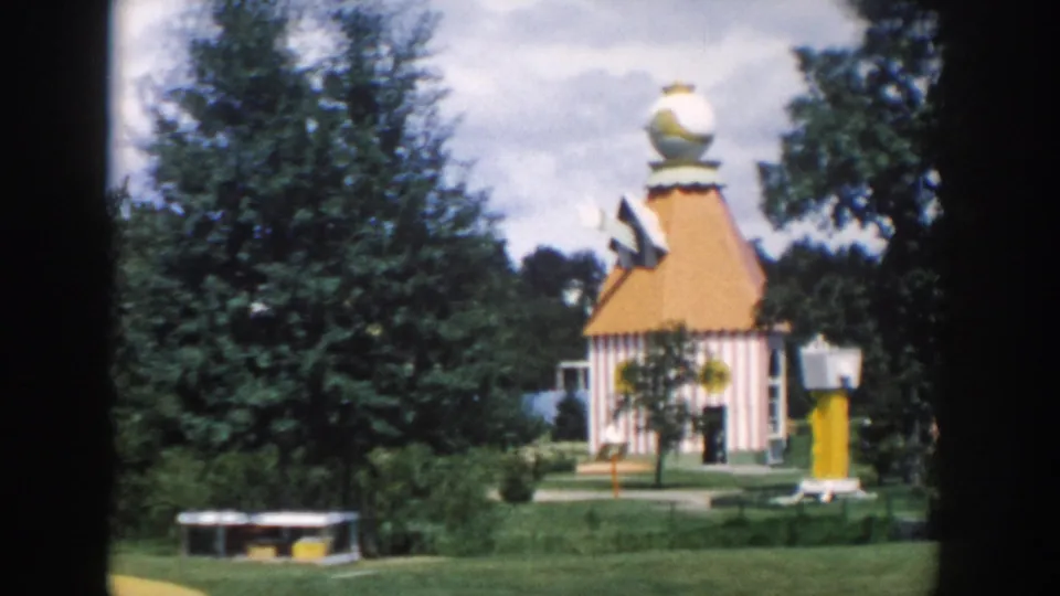 1954 Wisconsin Dells frame showing a colorful small building with a yellow roof, trees, lawns, and nearby park fixtures.