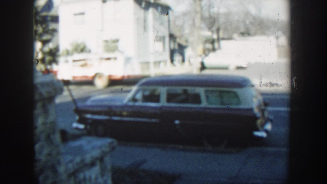 Soft 1954 Wisconsin Dells home-movie frame of a dark station wagon along a residential street with a truck and houses behind.