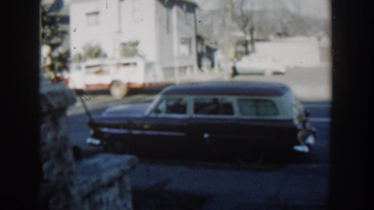 1954 Wisconsin Dells frame showing a side view of a dark station wagon filmed through foreground posts on a neighborhood street.