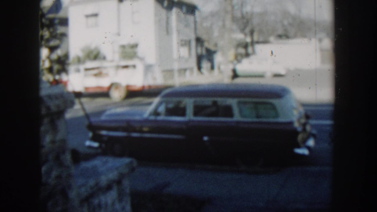 1954 Wisconsin Dells home-movie frame showing a dark station wagon parked outside a white house, viewed through a stone porch frame.