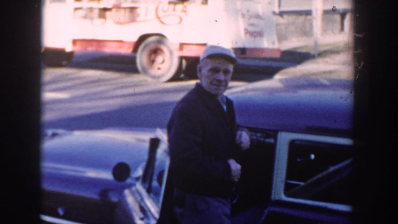 Older man in a cap stands beside a blue car with a delivery truck behind him.