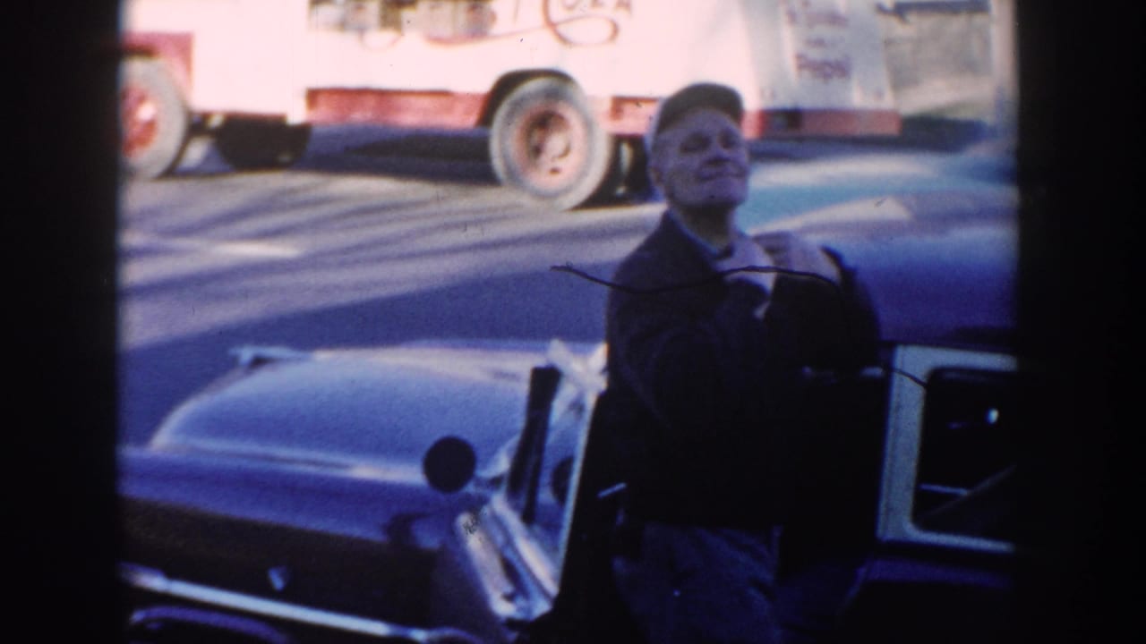 The older man appears to smile toward the camera while zipping or holding his jacket beside the blue car in the 1954 home movie.