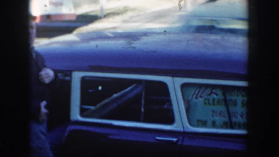 1954 Wisconsin Dells home-movie view of the parked blue station wagon, its rear-window sign, and a partial adult figure at frame edge.