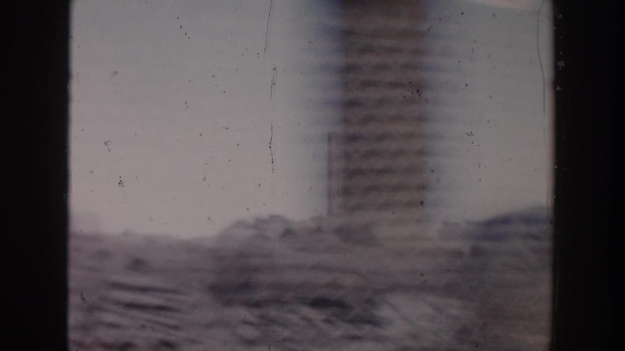 1956 Grand Rapids car-window view of a construction area with stacked masonry or beams and a tall soft-focused structure beyond.