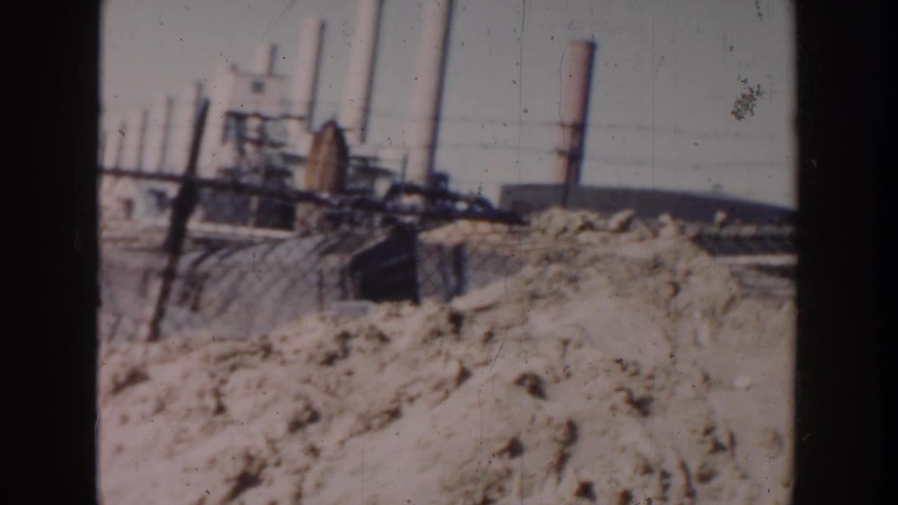 1956 Grand Rapids home-movie frame showing sandy rubble, rough fencing, and distant industrial smokestacks beyond the worksite.