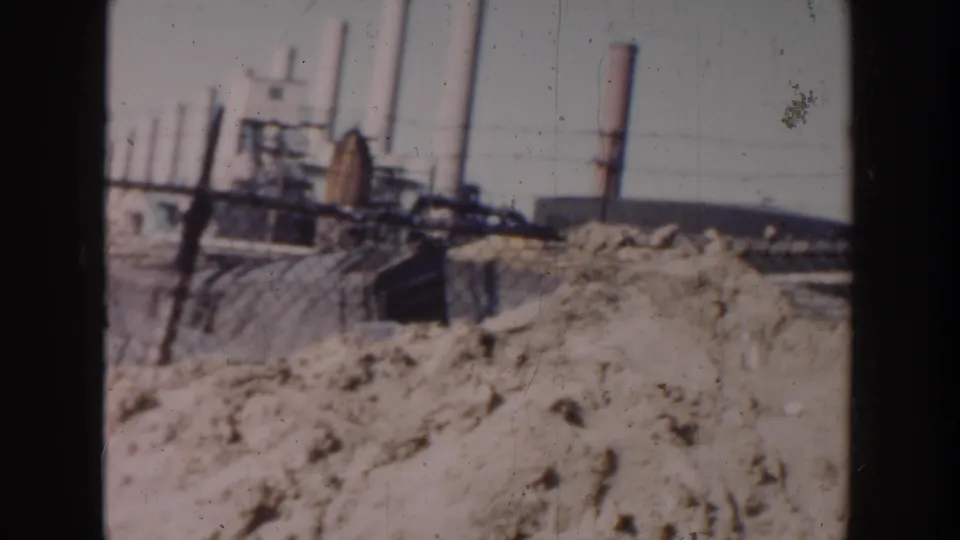 1956 Grand Rapids home-movie frame showing sandy rubble, rough fencing, and distant industrial smokestacks beyond the worksite.