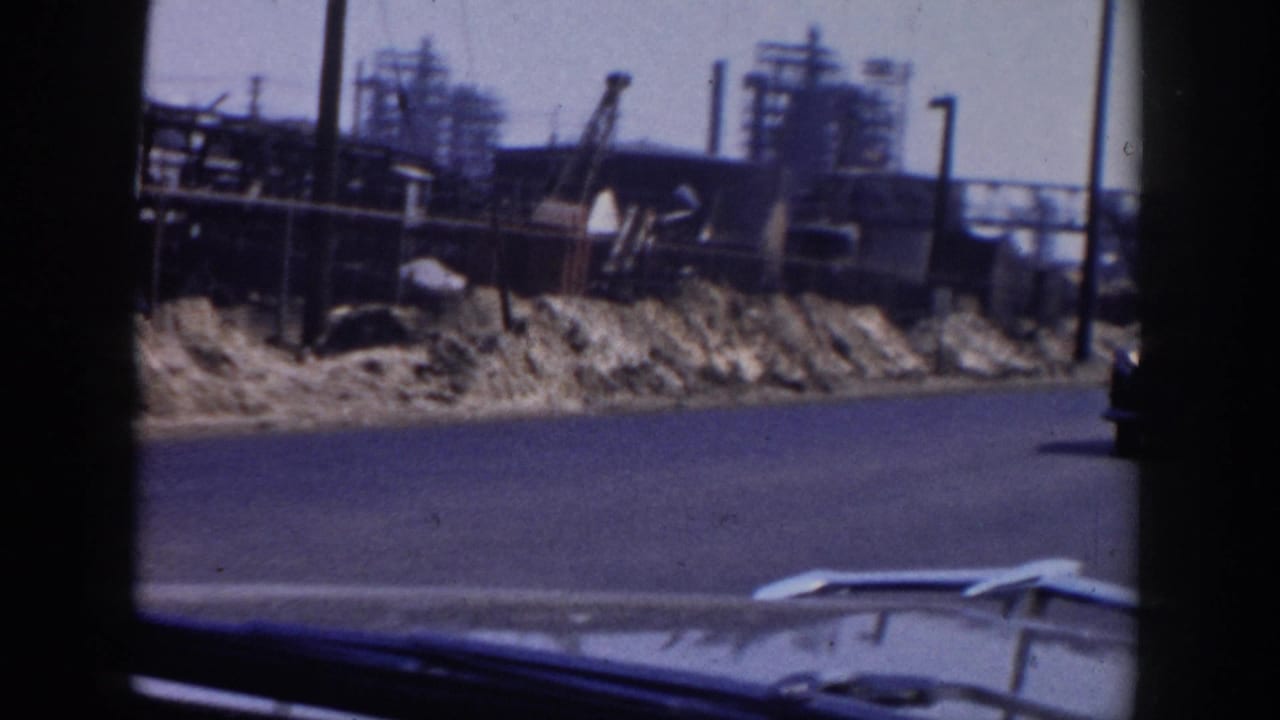 Roadside industrial area with gravel piles, cranes, pipework, and the edge of a car hood.