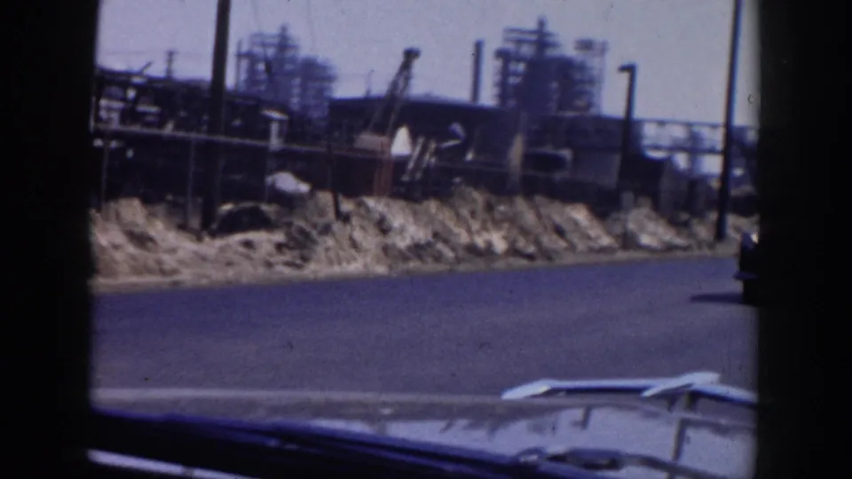 1956 Grand Rapids roadside view from a moving car, showing gravel piles, industrial structures, cranes, and part of the vehicle hood.