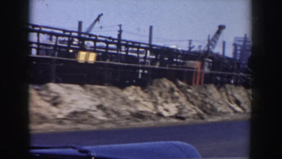 Passenger-side view of an industrial area in Grand Rapids, with pipe racks, cranes, gravel mounds, and a dark vehicle edge in frame.