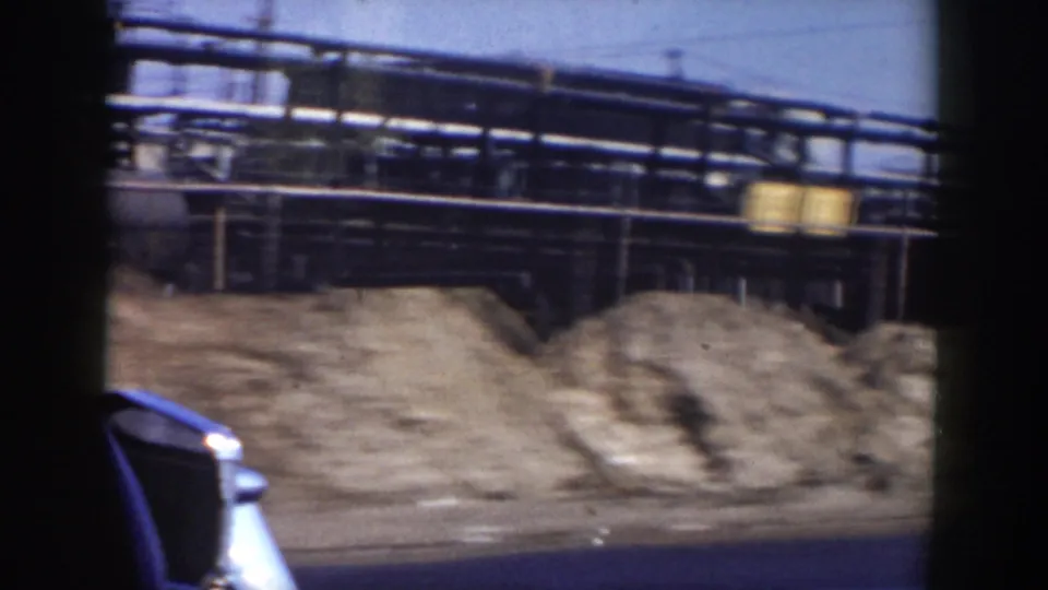 1950s driving view in Grand Rapids, showing a classic car, gravel piles, and industrial pipework behind a roadside fence or railing.