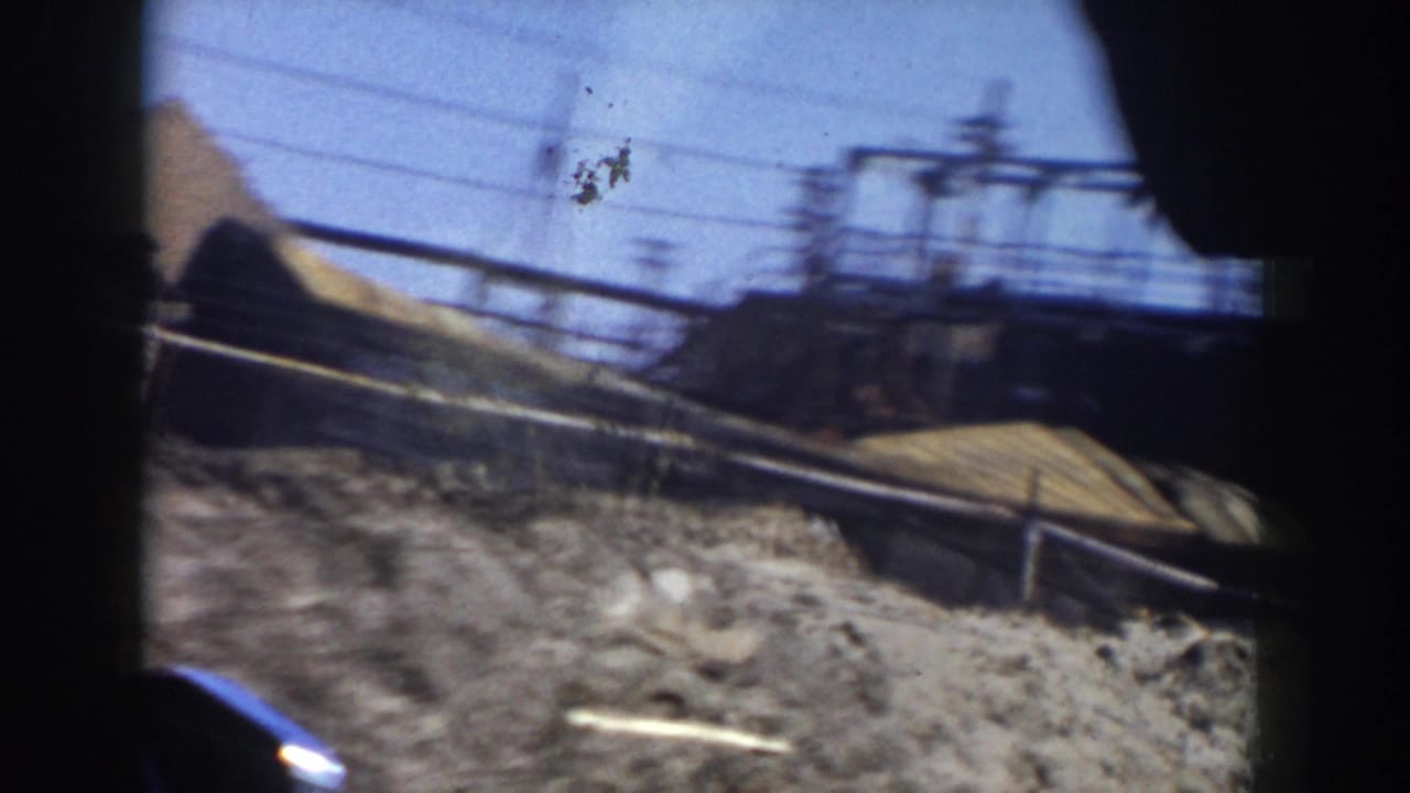 Blurred roadside construction area seen through a vehicle window with dirt, pipes, poles, and overhead wires.