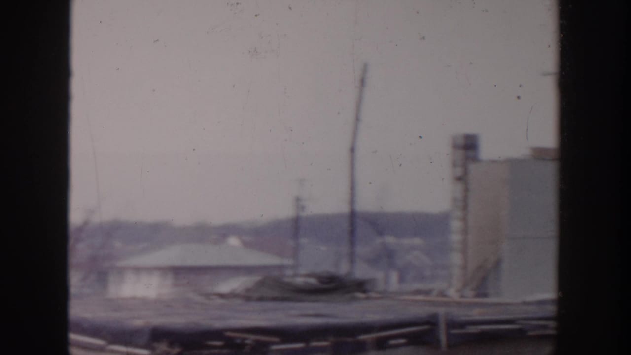 Rooftop view through a dark frame with pale structures, utility poles, and distant low buildings.