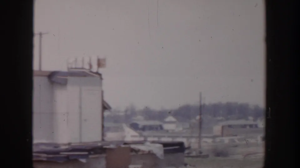 Grand Rapids 1956 home-movie frame with a partial rooftop structure, distant buildings, utility pole, and open ground.