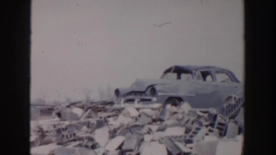 1956 Grand Rapids frame showing a wrecked mid-century car body on rubble with broken masonry and scattered debris.