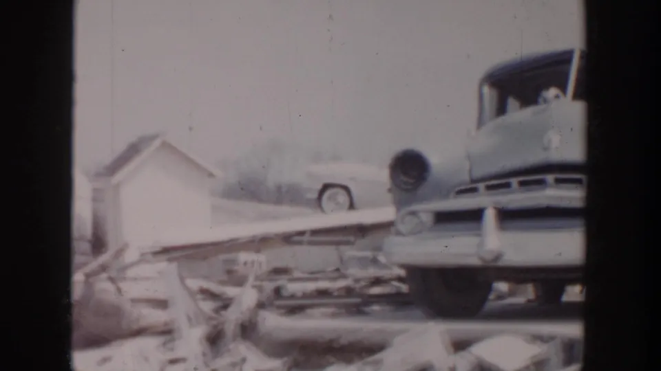 Close 1956 aftermath view with the front of a 1950s car, a trailer edge, and debris across the ground.