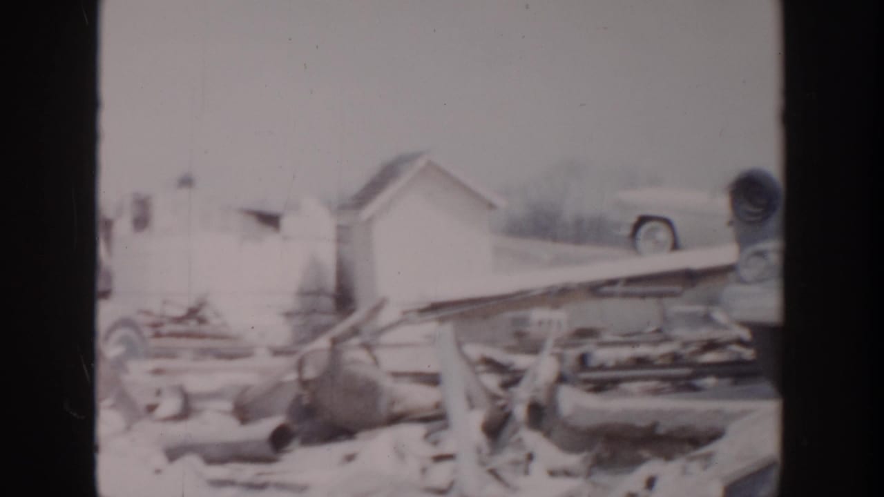 1956 Grand Rapids marina aftermath with snow, damaged dock material, a small shed, and a partly obscured 1950s automobile.