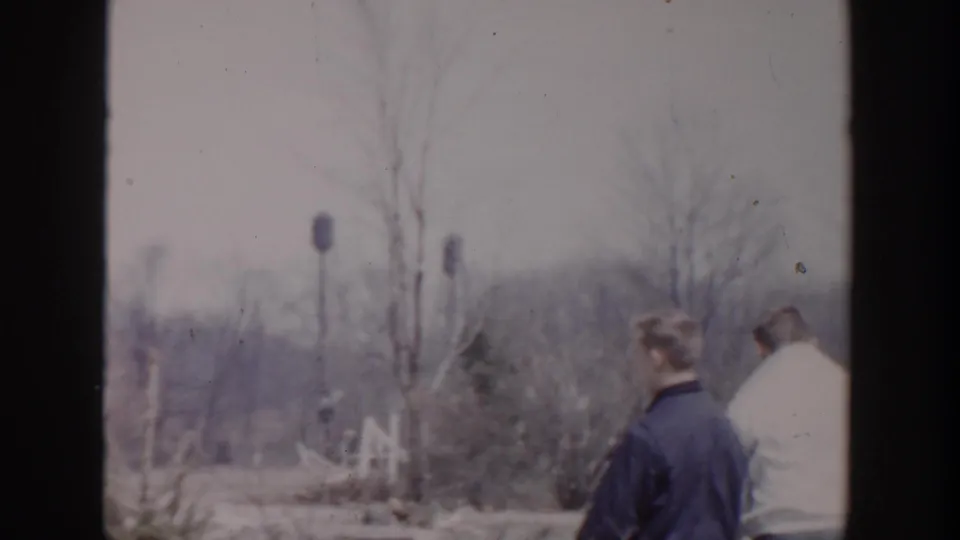 Home-movie frame from Grand Rapids in 1956 showing two people from behind beside bare trees and a quiet winter yard.