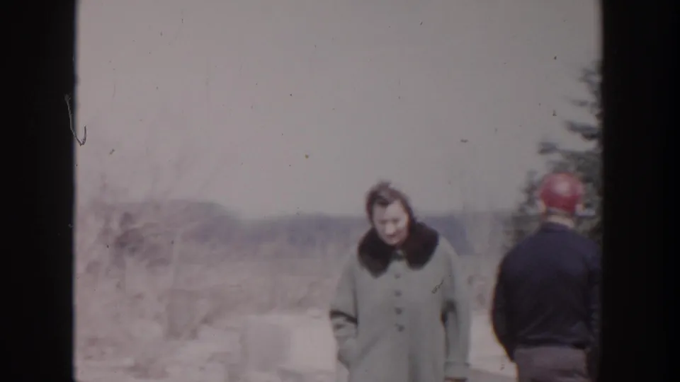 A woman in a light coat with a dark collar stands near a man in a red cap at the snowy Michigan house site.