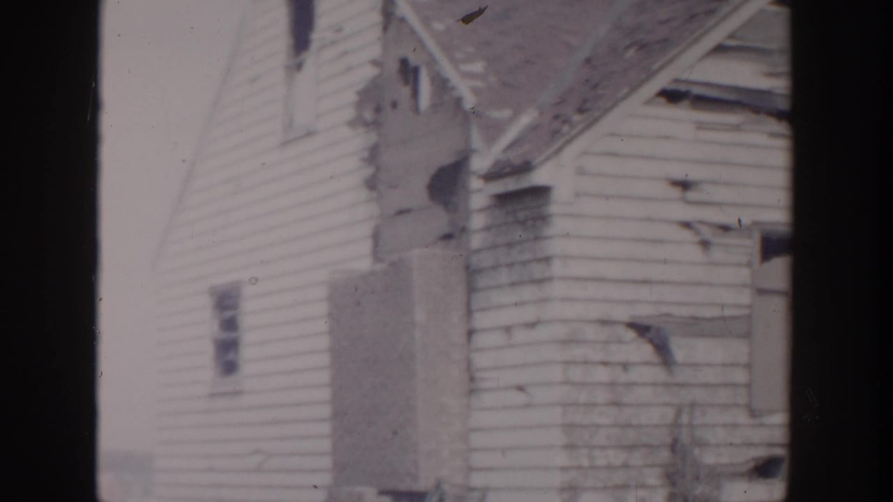 1956 Grand Rapids home-movie frame showing a damaged white clapboard house with torn siding and a compromised roofline.