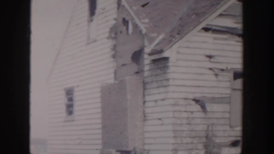 1956 Grand Rapids home-movie frame showing a damaged white clapboard house with torn siding and a compromised roofline.
