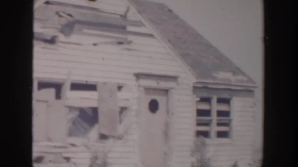 1956 Grand Rapids frame of a damaged clapboard house front, including broken windows, torn siding, and a door with an oval window.