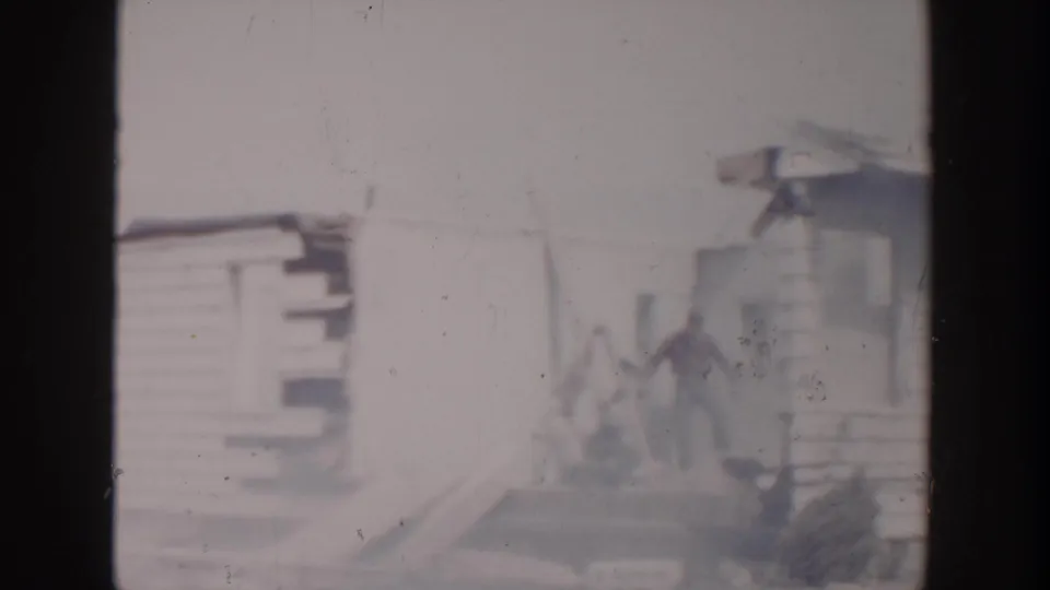 1956 Grand Rapids home-movie frame showing a weathered wooden house, damaged siding, and a distant person near the structure.
