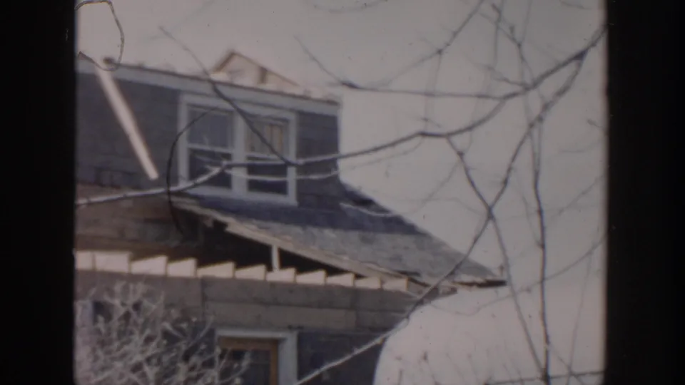 1956 Grand Rapids frame showing a damaged dormer roof, exposed framing, and bare branches in front of a weathered house.