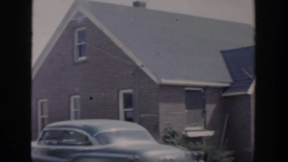1956 Grand Rapids home-movie frame showing a wooden residential house, steep roofline, and a parked mid-century car.