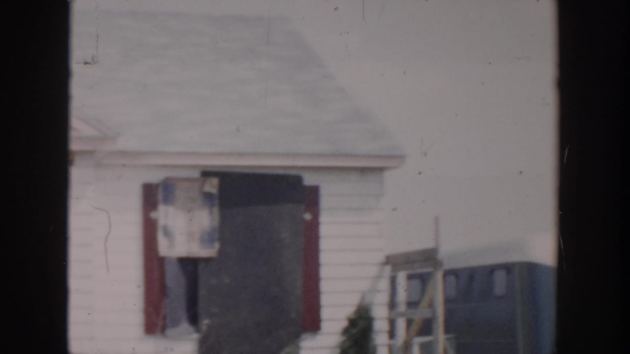 Grand Rapids 1956 home-movie frame showing a damaged white house with a dark doorway, broken window area, and red shutters.