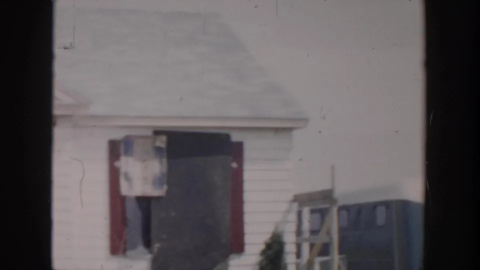 Grand Rapids 1956 home-movie frame showing a damaged white house with a dark doorway, broken window area, and red shutters.