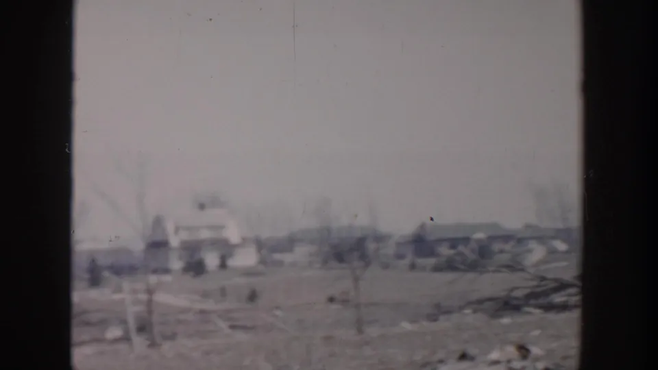1956 Grand Rapids home-movie frame showing a soft, distant view of leafless trees, low buildings, and debris-strewn ground.