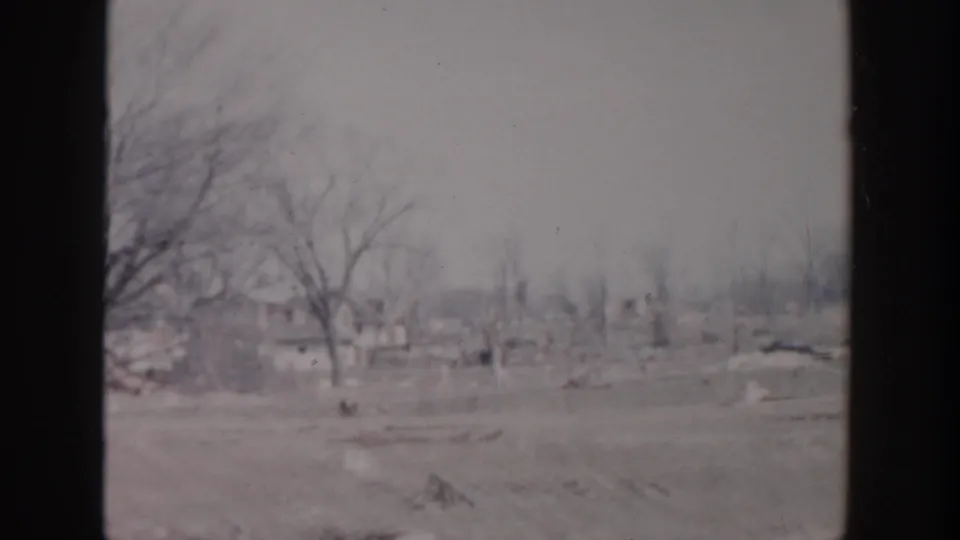 1956 Grand Rapids frame showing bare trees, disturbed ground, debris, and distant residential structures in a damaged outdoor landscape.
