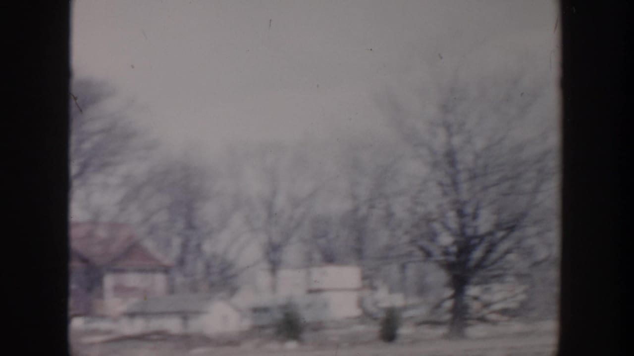 Grand Rapids 1956 home-movie frame showing detached houses and leafless trees along a subdued residential street.