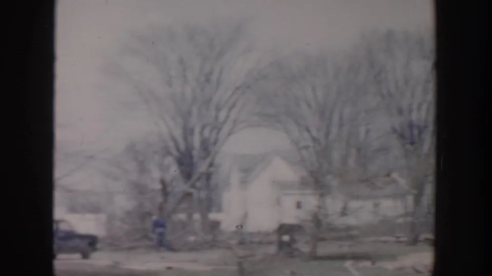 Grand Rapids 1956 frame showing a house, leafless trees, a parked car, and a small distant person on or near the residential street.