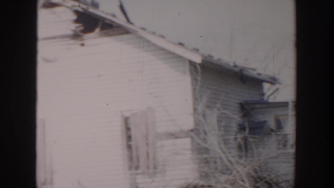 1956 Grand Rapids home-movie frame showing a white clapboard house corner with damaged roof edge, bare branches, and soft 8mm exposure.