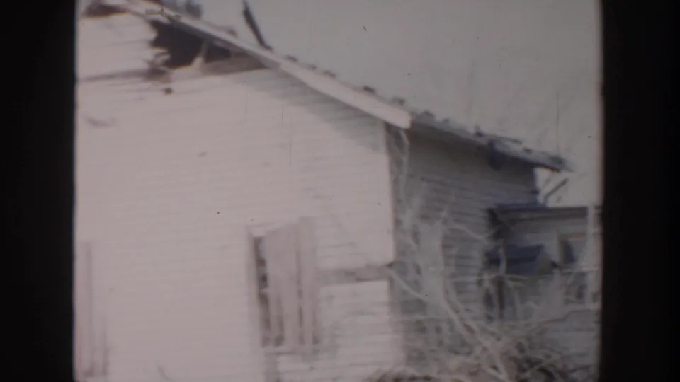 1956 Grand Rapids home-movie frame showing a white clapboard house corner with damaged roof edge, bare branches, and soft 8mm exposure.