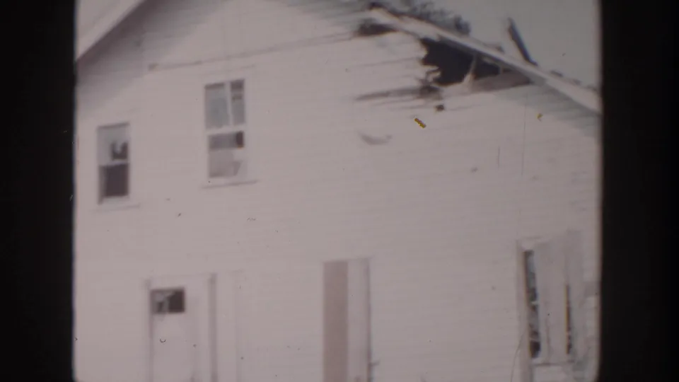 1956 Grand Rapids frame showing the side of a white clapboard house with upper windows, a side door, and visible roof-edge damage.