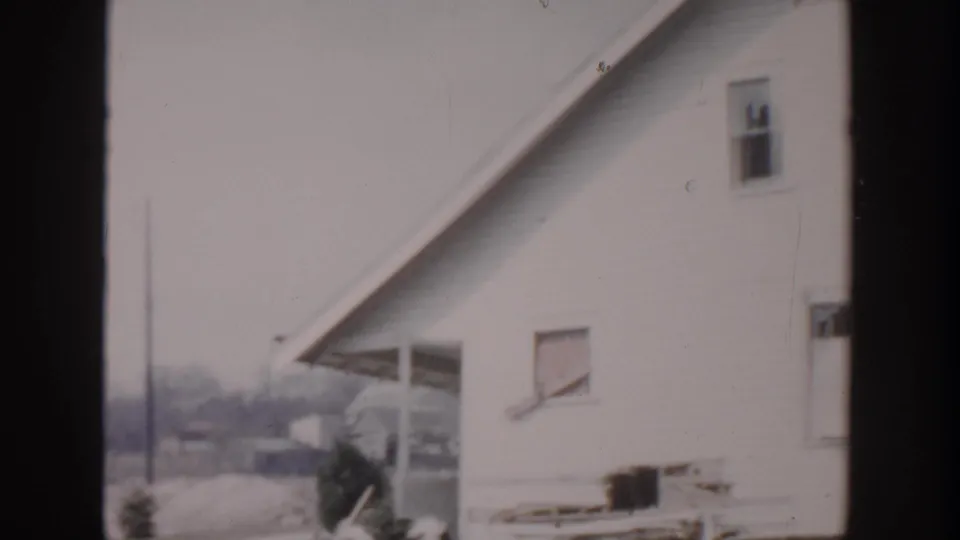 1956 Grand Rapids home-movie frame showing the white house side, sloped roofline, porch area, small windows, and yard beyond.