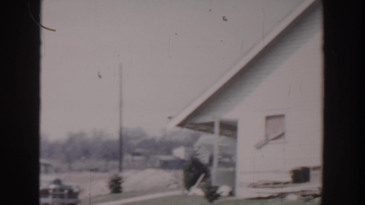 1956 Grand Rapids home-movie frame showing a white-sided residential exterior, driveway area, parked car, utility pole, and lawn.