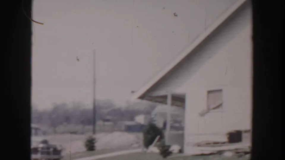 1956 Grand Rapids home-movie frame showing a white-sided residential exterior, driveway area, parked car, utility pole, and lawn.