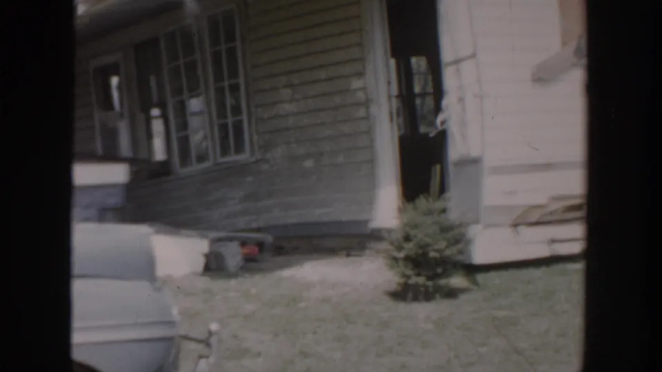 1956 Grand Rapids residential porch view with multi-pane windows, open doorway, lawn, small evergreen, and soft 8mm film wear.