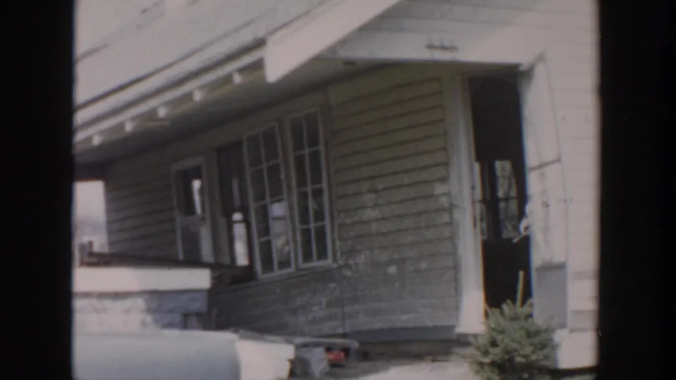 Soft 8mm frame of a Grand Rapids house porch in 1956, showing windows, doorway, roofline, siding, lawn, and shrub.