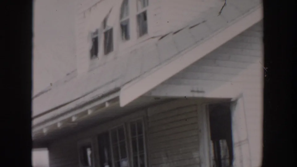 Tilted 1956 Grand Rapids home-movie frame showing house siding, upper windows, roofline, porch windows, and yard edge.