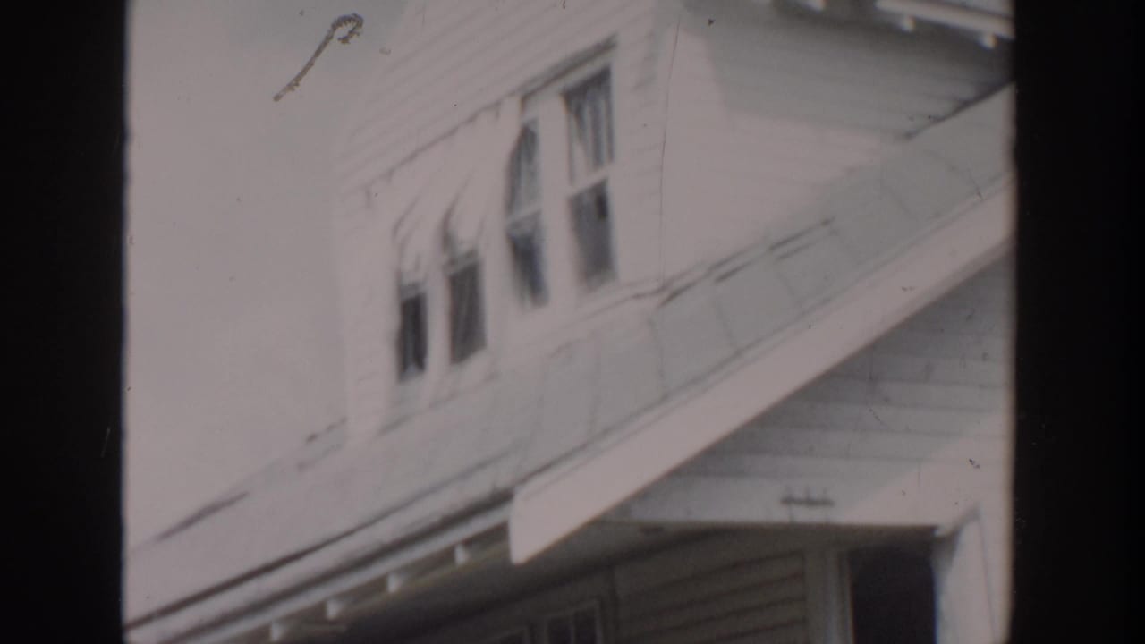 1956 Grand Rapids home-movie frame showing a white clapboard house with upper windows and damaged roofline detail.