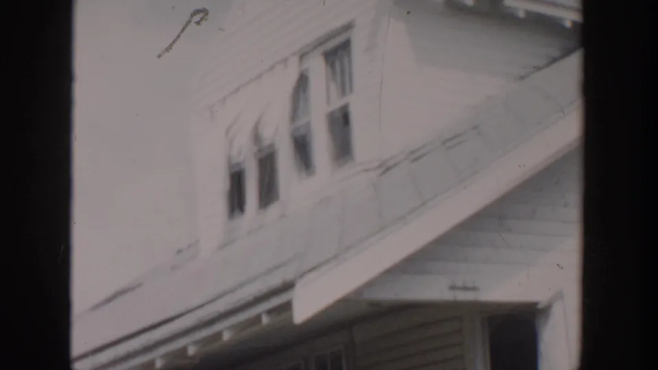 1956 Grand Rapids home-movie frame showing a white clapboard house with upper windows and damaged roofline detail.