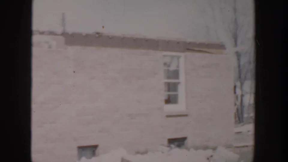 1956 Grand Rapids frame of a pale brick house wall in winter, showing a small window, snow at ground level, and bare branches at the edge.