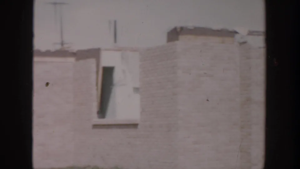 1956 Grand Rapids home-movie frame showing a brick house corner in winter with a dark window opening and weathered exterior details.