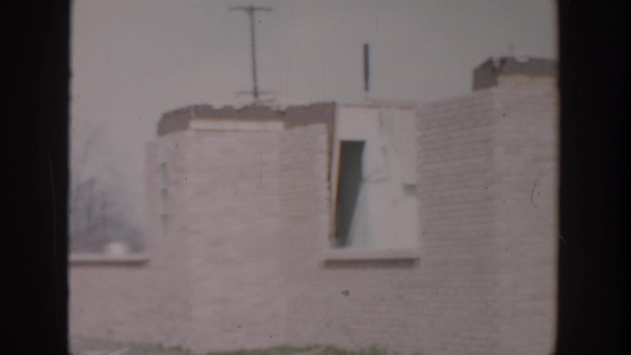 1956 Grand Rapids home-movie frame showing a pale brick wall, small window opening, roofline, and utility pole in the background.